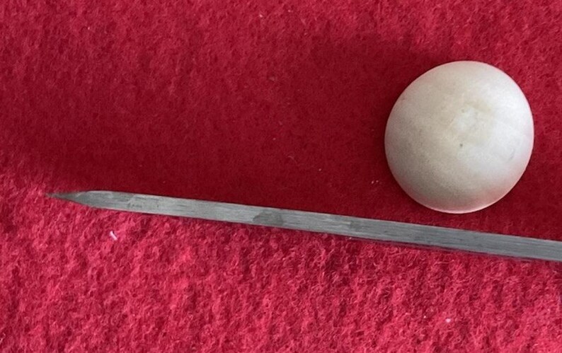 Puede incluir: Primer plano de un objeto esf&eacute;rico blanco y una herramienta met&aacute;lica larga y delgada con un extremo puntiagudo. Los objetos est&aacute;n sobre un fondo rojo.