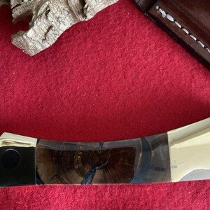 Puede incluir: Primer plano de una navaja de bolsillo con mango de madera y hoja plateada. El mango presenta un patr&oacute;n de vetas de madera oscura con detalles en negro y plata. La navaja est&aacute; parcialmente abierta, mostrando la hoja.