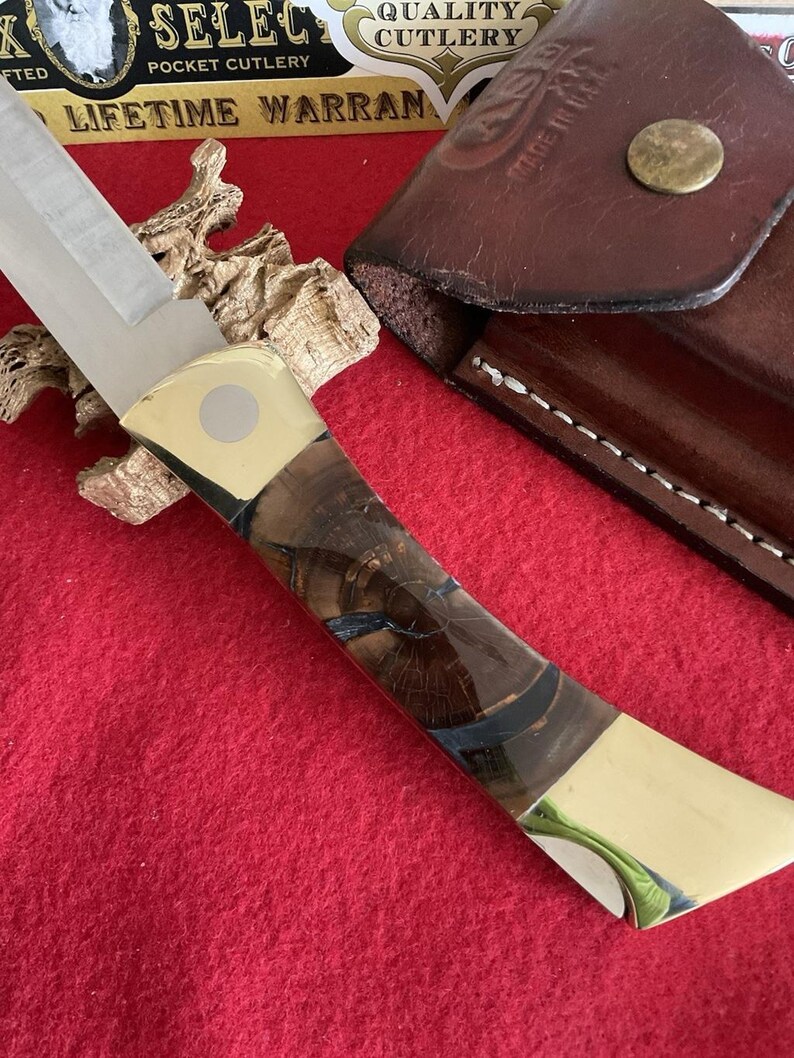Puede incluir: Una navaja de bolsillo con una hoja de acero pulido y un mango con un patr&oacute;n de vetas de madera con detalles azules. El mango tiene extremos color lat&oacute;n. Una funda de cuero marr&oacute;n es visible en el fondo.