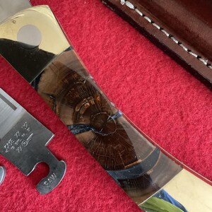 Puede incluir: Un cuchillo artesanal con un mango de madera con un patr&oacute;n de vetas &uacute;nico y detalles en lat&oacute;n. La hoja es parcialmente visible, junto con una pieza de metal separada. El cuchillo descansa sobre una superficie roja, con una funda de cuero en el fondo.
