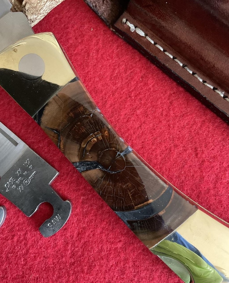 Puede incluir: Un cuchillo artesanal con un mango de madera con un patr&oacute;n de vetas &uacute;nico y detalles en lat&oacute;n. La hoja es parcialmente visible, junto con una pieza de metal separada. El cuchillo descansa sobre una superficie roja, con una funda de cuero en el fondo.