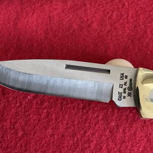 Puede incluir: Primer plano de una navaja de bolsillo Case XX USA. La hoja es plateada con un borde oscuro y el mango es dorado. El cuchillo est&aacute; sobre una superficie roja. El texto "CASE XX USA" es visible en la hoja.
