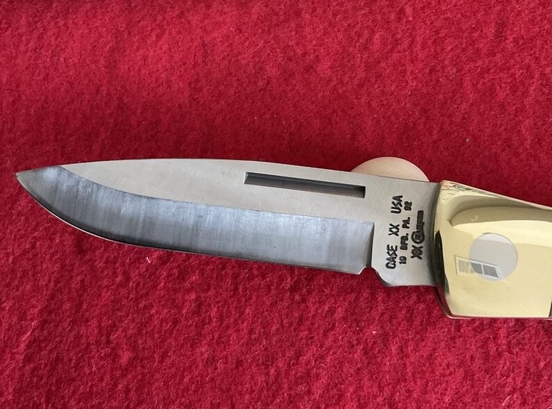 Puede incluir: Primer plano de una navaja de bolsillo Case XX USA. La hoja es plateada con un borde oscuro y el mango es dorado. El cuchillo est&aacute; sobre una superficie roja. El texto "CASE XX USA" es visible en la hoja.