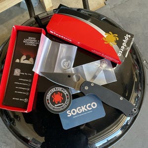 May include: A black folding knife with a silver blade, a red and black box, and a blue card with the text "SOGKCO". The knife is accompanied by a sticker that says "Crafted for Life" and "Spyderco".