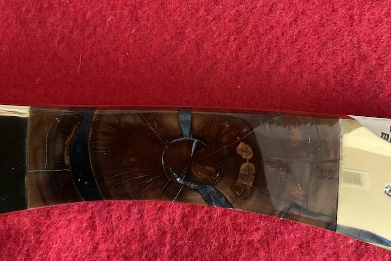 Puede incluir: Primer plano de un mango de cuchillo con un patr&oacute;n de vetas de madera marr&oacute;n oscuro. El mango est&aacute; acentuado con elementos negros y dorados. El cuchillo parece ser un art&iacute;culo hecho a mano, que muestra un dise&ntilde;o y una artesan&iacute;a &uacute;nicos.