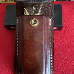Puede incluir: Una funda de cuchillo de cuero marr&oacute;n con cierre de bot&oacute;n a presi&oacute;n de lat&oacute;n. La funda est&aacute; cosida con hilo blanco y contiene una peque&ntilde;a navaja plegable. El cuchillo tiene un mango dorado y un llavero. El fondo incluye una superficie roja y una caja negra.