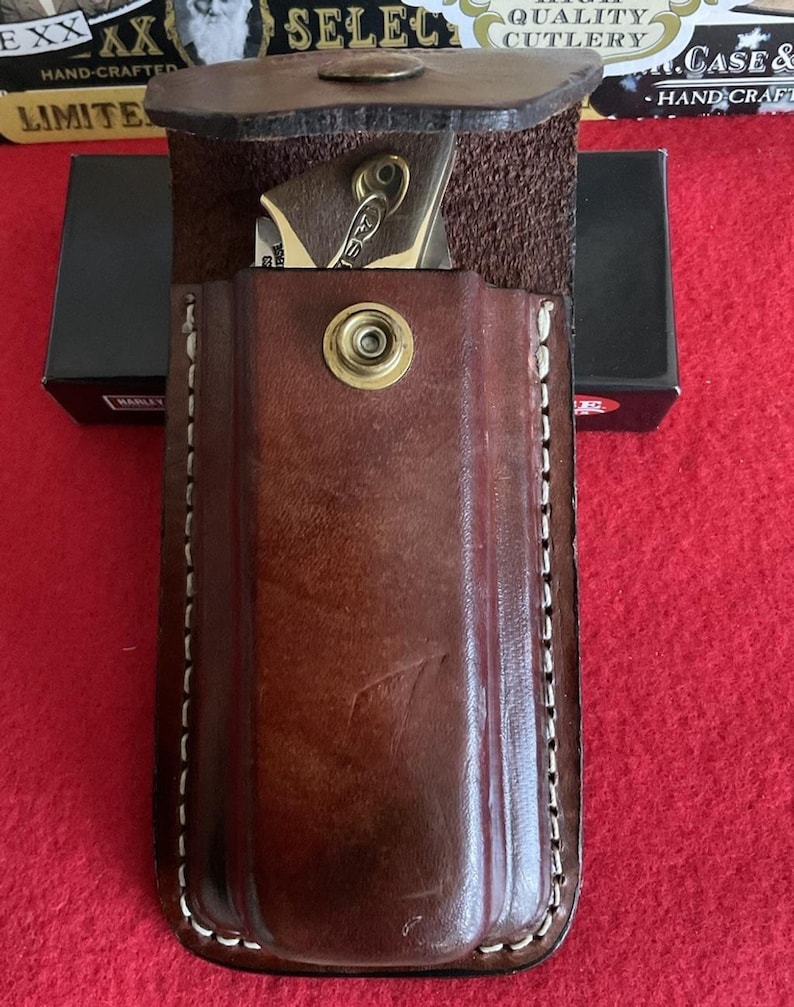 Puede incluir: Una funda de cuchillo de cuero marr&oacute;n con cierre de bot&oacute;n a presi&oacute;n de lat&oacute;n. La funda est&aacute; cosida con hilo blanco y contiene una peque&ntilde;a navaja plegable. El cuchillo tiene un mango dorado y un llavero. El fondo incluye una superficie roja y una caja negra.