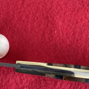 Puede incluir: Una bola de madera y un cuchillo con un mango de madera estampada y una hoja plateada se muestran sobre un fondo rojo. El mango del cuchillo presenta una mezcla de tonos de madera oscuros y claros.