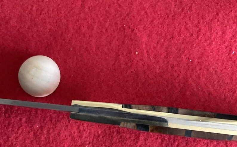 Puede incluir: Una bola de madera y un cuchillo con un mango de madera estampada y una hoja plateada se muestran sobre un fondo rojo. El mango del cuchillo presenta una mezcla de tonos de madera oscuros y claros.