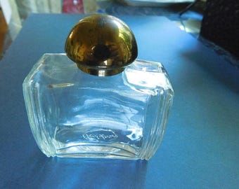 May include: A clear, octagonal glass perfume bottle with a gold-coloured, mushroom-shaped stopper. The bottle has a slightly curved design and a small, oval-shaped marking near the base. The bottle sits on a blue surface.