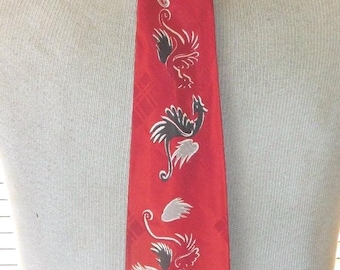 May include: A bright red necktie featuring a repeating pattern of stylized winged dragon figures in alternating shades of silver and black. The fabric has a subtle, tonal plaid texture throughout, and the dragons are arranged vertically.