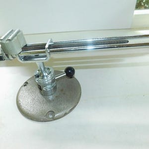 May include: A silver-colored metal vise with a long, curved handle. The vise has a round base and a clamp at the end of the handle. The handle is approximately 12 inches long.
