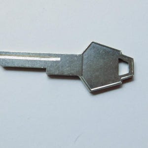 Puede incluir: Una llave plateada con un cuerpo rectangular y una cabeza geométrica. La llave tiene una sola ranura y un pequeño agujero en la cabeza. La llave está diseñada para una puerta o cerradura.