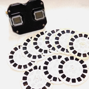 May include: A black plastic vintage View-Master viewer with a set of 10 white plastic reels. Each reel has 14 circular images. The reels have text on them, including "Wonder Book" and "Story Book".