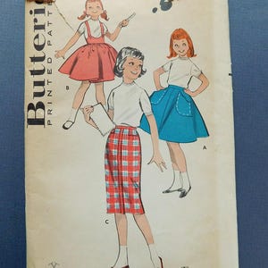Puede incluir: Sobre de patrones impresos Butterick vintage con ilustraciones de niñas con diferentes atuendos. Los atuendos incluyen una falda roja con tirantes, una falda azul con bolsillos y una falda a cuadros. El sobre está desgastado.