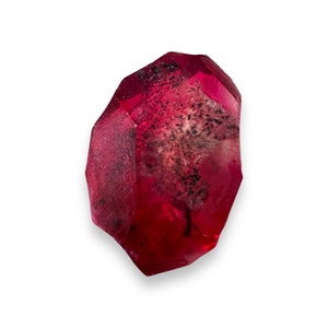 May include: A faceted, translucent, red crystal with dark inclusions. The gemstone has a polished, glossy appearance and is set against a white background. The crystal's shape is irregular, with multiple facets.