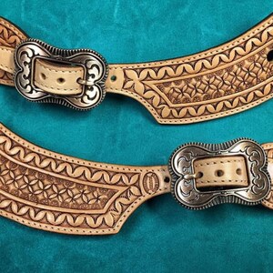 May include: Two light tan leather spur straps with intricate floral and geometric tooling. Each strap features a silver-colored buckle and a matching leather keeper. The straps are displayed on a teal background.