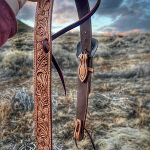 May include: A handcrafted leather horse bridle with intricate floral tooling. The bridle features a light brown, carved leather strap with a dark brown leather strap and accents. The background shows a blurred outdoor scene.