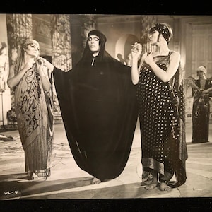 May include: Three women in a black and white photo wearing long, flowing robes with intricate patterns. The woman in the center is wearing a black robe with a hood. The women are standing in a room with a marble floor and ornate columns.