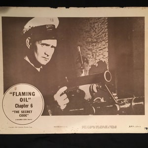 Op de afbeelding: Een zwart-witfoto van een man in een matrozenpet die een pistool vasthoudt. De foto is van de film "Flaming Oil" Hoofdstuk 6 "De geheime code".