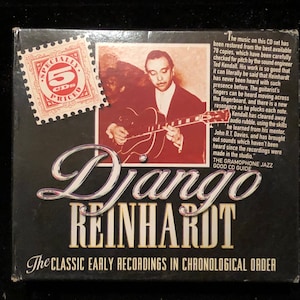 Peut inclure: Un coffret de 5 CD noir et blanc avec une photo de Django Reinhardt jouant de la guitare. La boîte porte le texte "Django Reinhardt" en grandes lettres blanches et "The Classic Early Recordings in Chronological Order" en lettres blanches plus petites. La boîte porte également un timbre rouge avec le texte "5 CD" et "Prix spécial".