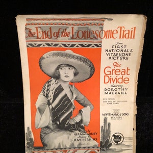 Puede incluir: Una portada de partitura para la canción "The End of the Lonesome Trail" de la película First National & Vitaphone "The Great Divide" protagonizada por Dorothy Mackaill. La portada presenta a una mujer con un sombrero y una serapa, sentada en una alfombra con un cactus en el fondo.