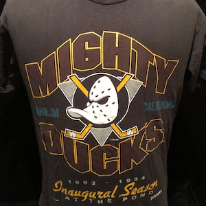 Puede incluir: Una camiseta negra con un gráfico amarillo y marrón de un pato jugador de hockey con el texto "Mighty Ducks" y "Inaugural Season 1993-1994 At The Pond".