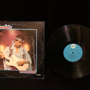 May include: A black vinyl record album cover featuring Jimi Hendrix in concert. The cover has a photo of Jimi Hendrix playing a white electric guitar. The album title is "In Concert" and is printed in red letters.