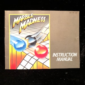 May include: A vintage instruction manual for the game "Marble Madness". The cover features a colorful illustration of three marbles rolling down a track.