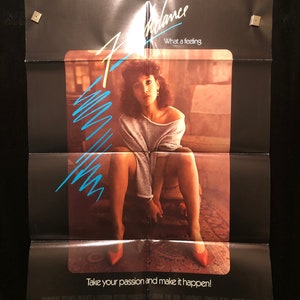 May include: A movie poster for the film "Flashdance" featuring Jennifer Beals in a grey sweater and red shoes. The poster has the text "Flashdance" in a blue and white font and the tagline "What a feeling." The poster also has the text "Take your passion and make it happen!" at the bottom.