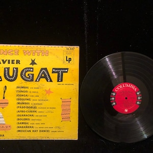 May include: A yellow vinyl record cover with a black and white image of Xavier Cugat and his orchestra. The cover features the text "Dance With Xavier Cugat" and a list of songs with their corresponding dance styles, including rumba, tango, conga, beguine, mambo, paso-doble, afro-cuban, guaracha, bolero, samba, habanera, and Mexican Hat Dance.