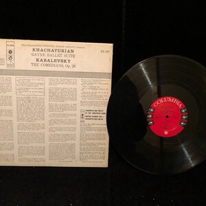 May include: A black vinyl record with a red label. The label has the text "Columbia" and "The Comedians, Op. 26" printed on it. The record is part of a set that includes a record sleeve with the text "Khachaturian Gayne Ballet Suite Kabalevsky The Comedians, Op. 26" printed on it.