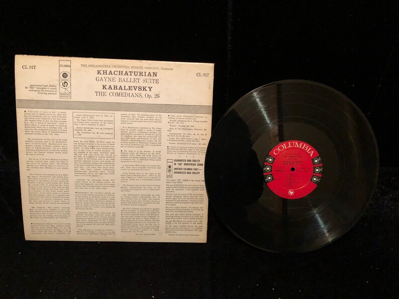 May include: A black vinyl record with a red label. The label has the text "Columbia" and "The Comedians, Op. 26" printed on it. The record is part of a set that includes a record sleeve with the text "Khachaturian Gayne Ballet Suite Kabalevsky The Comedians, Op. 26" printed on it.