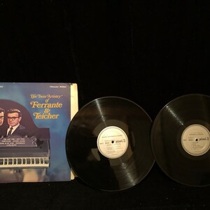 以下が含まれることがあります： 「The Twin Artistry of Ferrante & Teicher」のヴィンテージビニールレコードアルバムのカバー。スーツとメガネをかけた2人の男性が特徴です。2枚の黒いビニールレコードがアルバムカバーの隣にあります。アルバムカバーは青と白です。