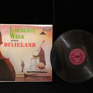 Puede incluir: Una portada de álbum de vinilo vintage con Lawrence Welk tocando música Dixieland. La portada presenta una camisa a rayas rojas y blancas, un banjo y una variedad de instrumentos de viento. El álbum está en el sello Coral.