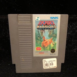 May include: A gray Nintendo Entertainment System (NES) game cartridge for the game Ikari Warriors. The cartridge has a label with the game title and a picture of the game's characters. The label also includes the text "SNK" and "Licensed by Nintendo for play on the NES."