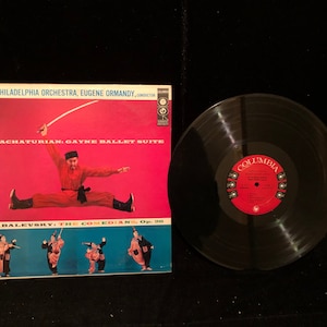 May include: A vintage vinyl record album cover featuring a red background with a man in a red and black costume performing a split. The album title is "KHACHATURIAN: GAYNE BALLET SUITE" and the album artist is "THE PHILADELPHIA ORCHESTRA, EUGENE ORMANDY, CONDUCTOR". The album also features a second piece by "KABALEVSKY: THE COMEDIANS, Op. 20".