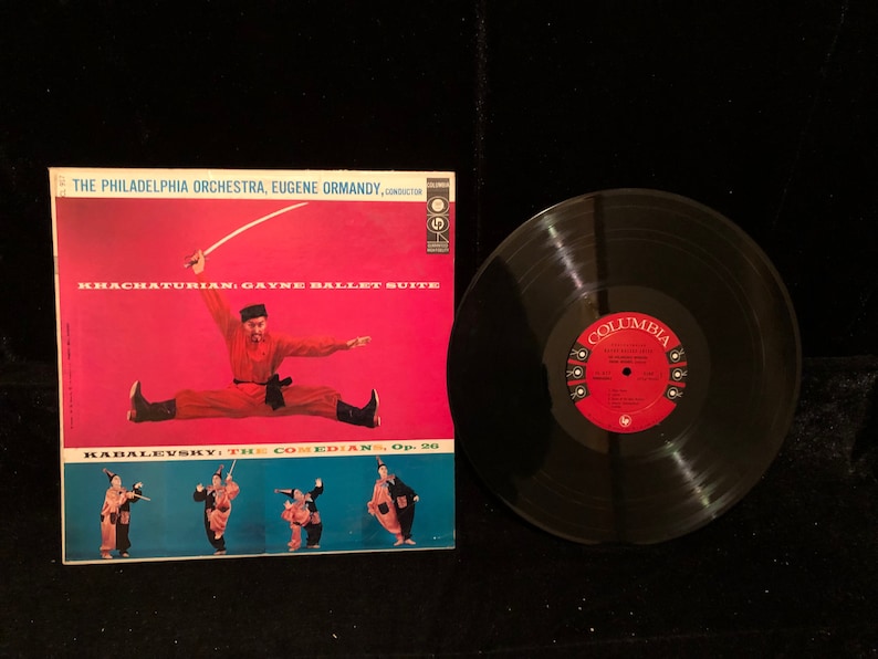 May include: A vintage vinyl record album cover featuring a red background with a man in a red and black costume performing a split. The album title is "KHACHATURIAN: GAYNE BALLET SUITE" and the album artist is "THE PHILADELPHIA ORCHESTRA, EUGENE ORMANDY, CONDUCTOR". The album also features a second piece by "KABALEVSKY: THE COMEDIANS, Op. 20".