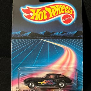 Könnte beinhalten: Ein schwarzes Hot Wheels Split Window '63 Modellauto aus Druckgussmetall auf der Originalverpackung. Die Verpackung hat einen blau-violetten Hintergrund mit dem Hot Wheels-Logo und dem Text "FOR AGES OVER 3." Das Auto hat Flammenaufkleber.