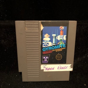 May include: A gray Nintendo Entertainment System game cartridge for Gyromite. The label features a pixelated image of a character jumping over a series of platforms. The text "Gyromite" is printed in white with a blue outline. The label also includes the Nintendo logo and a seal of quality.