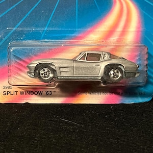 May include: Silver die-cast model of a Split Window '63 Corvette, packaged in a clear plastic case. The car has black wheels and a red interior. The packaging has the number 3985 and the text "SPLIT WINDOW '63".