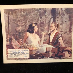 May include: A vintage movie poster for the film "Solomon and Sheba" starring Yul Brynner and Gina Lollobrigida. The poster features a scene of the two actors in a desert setting, with a backdrop of rocky cliffs and lush greenery.