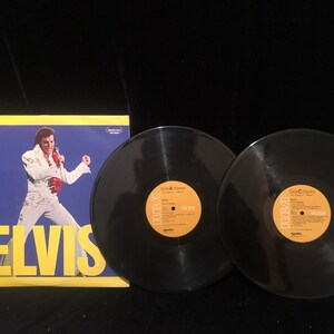 Peut inclure: Un coffret double vinyle d'Elvis Presley. La pochette de l'album présente une photo d'Elvis en combinaison blanche, chantant dans un microphone. Les étiquettes des disques sont jaunes avec du texte noir et le logo RCA Victor.