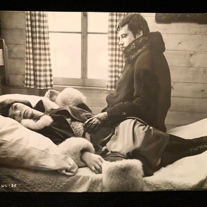 Pode incluir: Uma fotografia a preto e branco de um casal num quarto. A mulher está deitada numa cama com um casaco de pele, enquanto o homem está sentado na beira da cama, olhando para outro lado.