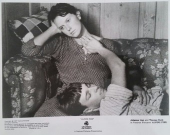 May include: Black and white photo of a young woman and man sitting on a couch. The woman is sitting upright with her arm resting on the back of the couch. The man is lying down with his head resting on the woman's lap. The photo is from the movie "Alpine Fire" starring Johanna Lier and Thomas Nock.