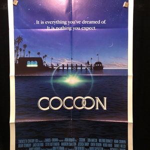 May include: A movie poster for the film "Cocoon" featuring a blue and purple sky with stars and a crescent moon. The title "Cocoon" is in white letters against a dark blue background. The poster also features a group of people on a dock, a boat, and a glowing orb in the water.