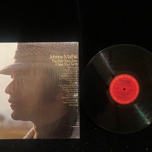 May include: A vinyl record album by Johnny Mathis, titled "The First Time Ever I Saw Your Face." The album cover features a profile of a person wearing a hat. A black vinyl record is next to the album.