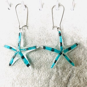 May include: A pair of starfish earrings. Each earring has a clear, five-pointed starfish design with turquoise blue and black accents. The earrings hang from silver-coloured ear wires, creating a beachy, nautical theme.