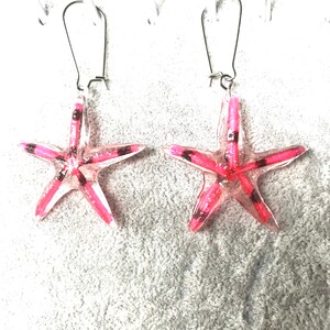 May include: A pair of starfish earrings. Each earring has a translucent pink starfish design with white and black detailing. The earrings are suspended from silver-coloured fish hook ear wires.