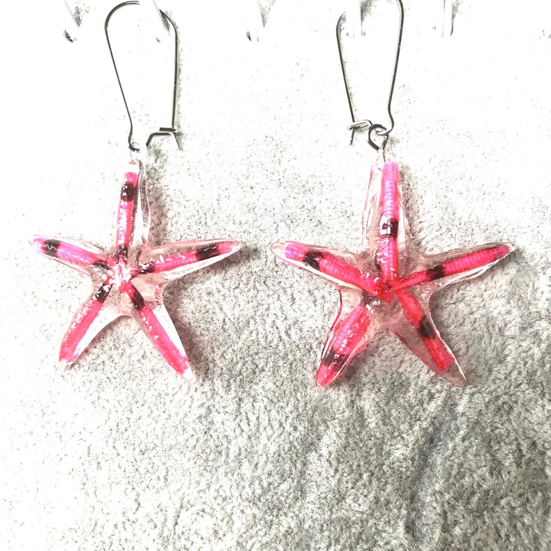 May include: A pair of starfish earrings. Each earring has a translucent pink starfish design with white and black detailing. The earrings are suspended from silver-coloured fish hook ear wires.